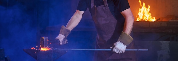 Professional mature blacksmith in safety apron and gloves beating on molten metal with hammer for shaping it Handwork and blacksmithing concept Close up