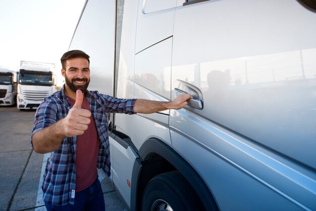 Профессиональный бородатый дальнобойщик средних лет стоит у своего грузовика-полуприцепа и держит палец вверх