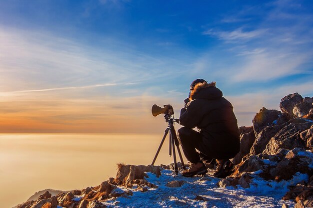 Professional photographer takes photos with camera on tripod on rocky peak at sunset. Dark tone