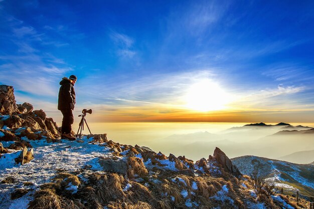 전문 사진 작가가 일몰 바위 봉우리에 삼각대에 카메라로 사진을 찍습니다.