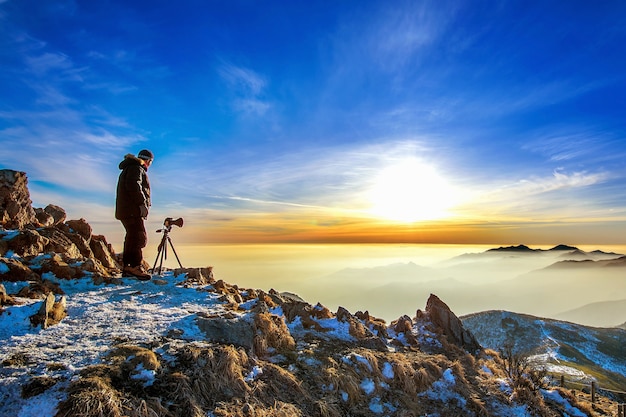 Professional photographer takes photos with camera on tripod on rocky peak at sunset