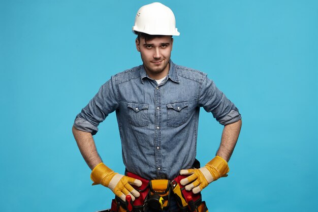 Professional skilled young male electrician in protective helmet and gloves holding hands on his waist