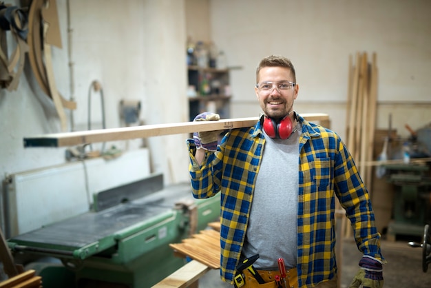 Professional smiling middle aged carpenter holding wood plank in woodworking workshop