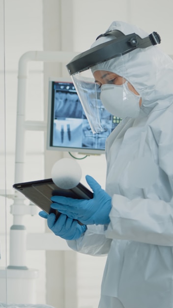 Professional stomatologists using digital tablet in dental cabinet at clinic. Dentist and assistant with ppe suits discussing patient oral care for operation and teeth surgery