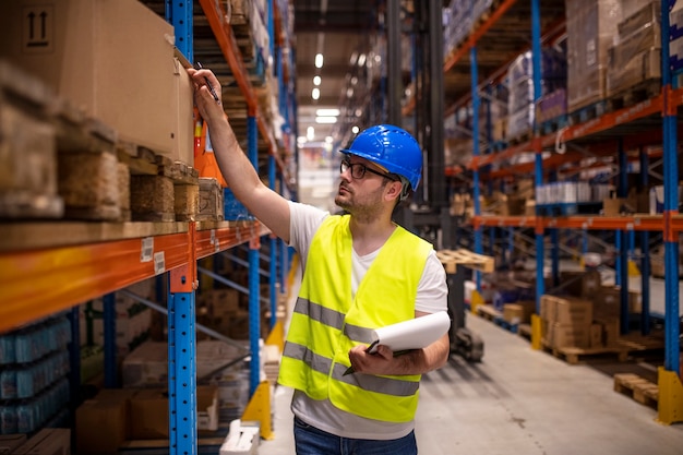 Professional warehouse worker in protective work wear holding checklist and checking inventory in storage room