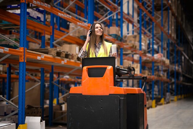 Professional warehouse worker with headset communication equipment driving forklift and relocating packages in storage center