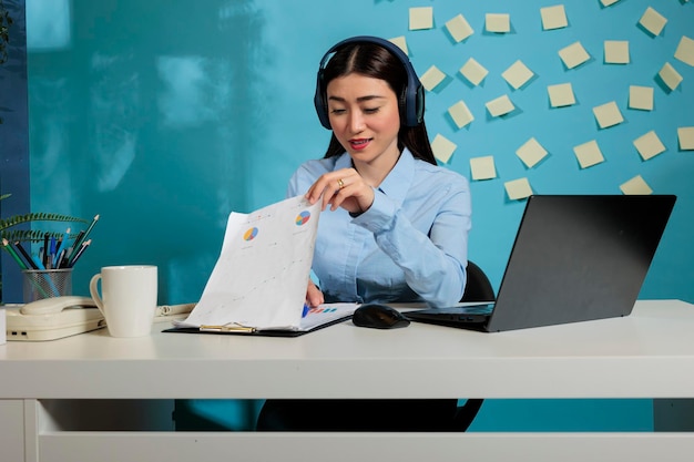 Professional woman listening to music with wireless headphones in office while checking monthly company statistics. Female employee looking at paperwork at workstation with laptop on desk.