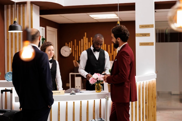 Professionals greeted by hotel staff at front desk, businessmen in suits arriving at luxury resort. People travelling for work partnership, doing check in procedure at reception.