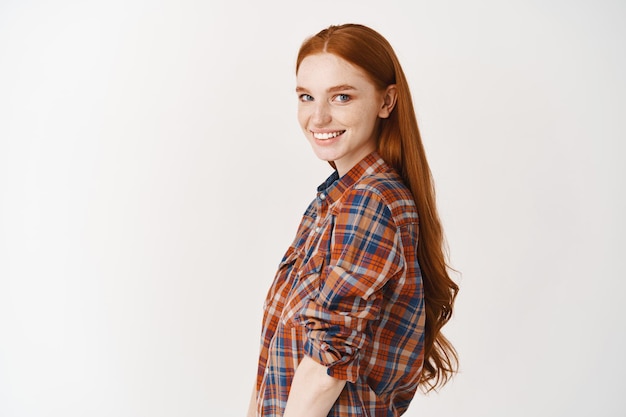 빨간 머리를 한 아름다운 10대 여성의 프로필, 머리를 앞으로 돌리고 웃고, 흰 벽 위에 서 있는