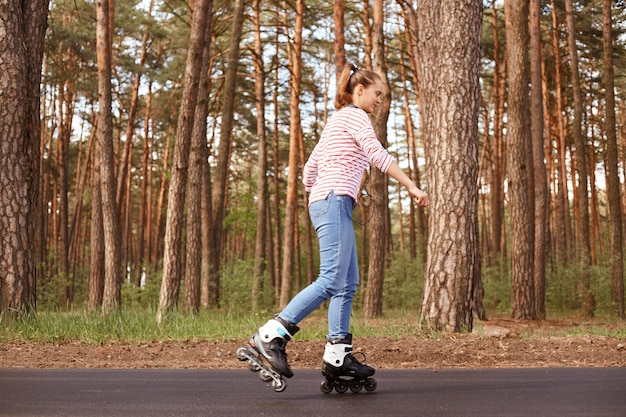 経験豊富な熟練したアクティブな若い女性が喜んでローラースケートをし、森の近くの道を歩き、健康的なライフスタイルにこだわり、ヘッドフォンを持ち、ストライプのスウェットシャツとジーンズを身に着けています。