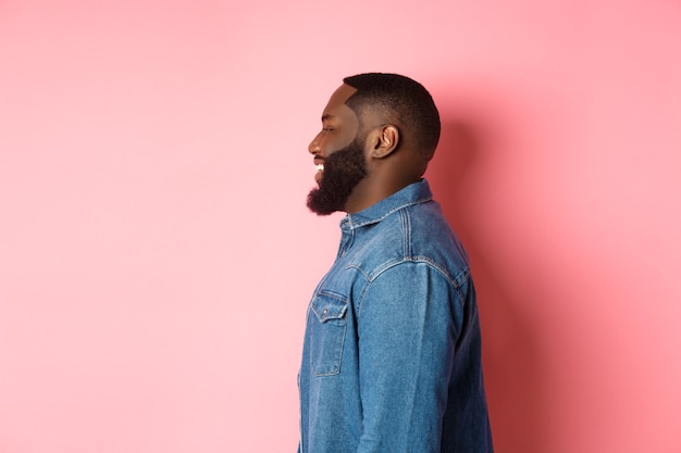 Profile of handsome bearded Black guy standing over pink background, smiling and looking left at copy space.