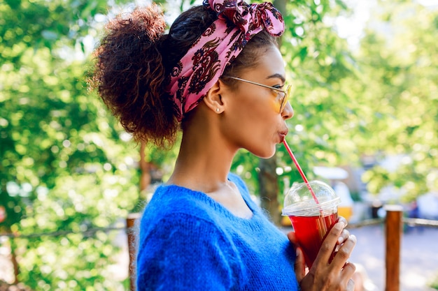 Profile of  happy  black  female  with stylish headband looking on horizon  and  holding cherry lemonade on hands