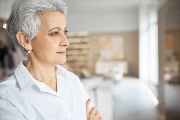 Profile picture of elegant stylish mature female entrepreneur wearing white formal shirt standing in modern office interior