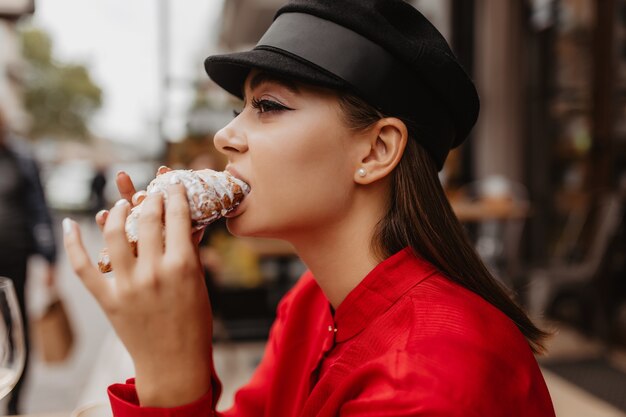 ベレー帽の茶色の髪のかわいい女の子のプロフィール写真。ストリートカフェで有名なアメリカのデザートの味を楽しむ女性