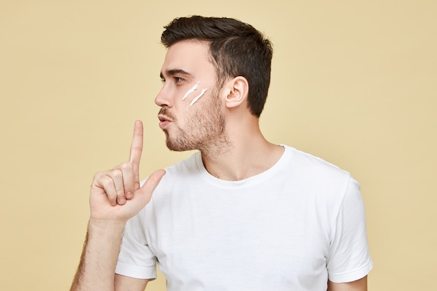 Profile portrait of attactive macho man with stubble and black hair posing  holding hand at his lips and blowing at index finger as if using psitol, having confident facial expression
