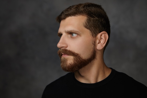 Profile portrait of attractive macho man with neat trimmed beard and mustache posing isolated at blank wall, frowning, expressing suspicion, looking away, having strict serious expression