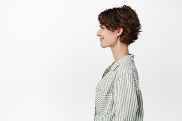 Profile portrait of beautiful smiling woman with short pixie hairstyle, looking left with happy smile, casual friendly face expression, standing in casual shirt over white background.