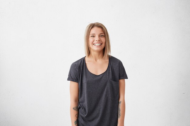 Profile portrait of beautiful young female with bobbed fair hair wearing casual grey T-shirt having tattoos on hands smiling pleasantly. Trendy hipster woman having good mood while posing