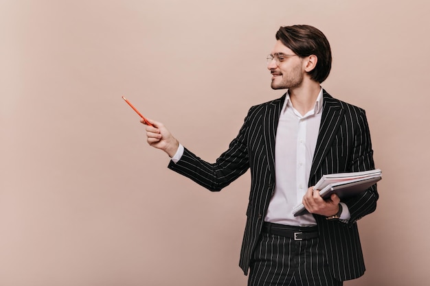 Free photo profile portrait of charming young brunette in elegant outfit holding notebooks in hand and pointing on free space aside with pencil man posing isolated over beige background