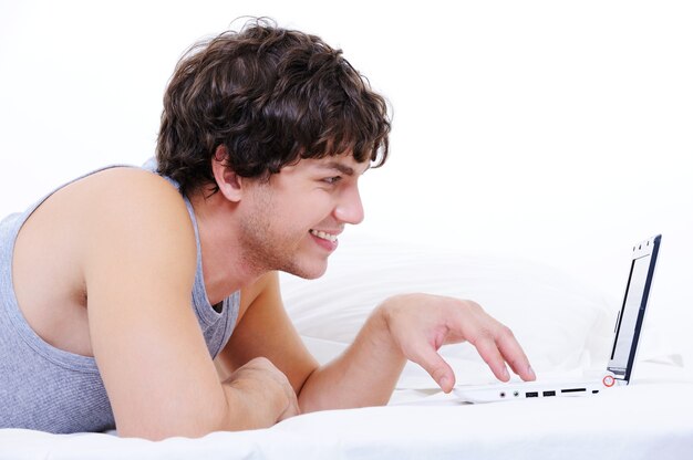 Profile portrait of a cheerful young male typing on laptop lying in his bed