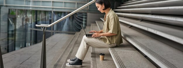 無料写真 ラップトップ女子学生を持つ若いアジア人女性の横顔のポートレートは建物の外の階段に座っており、