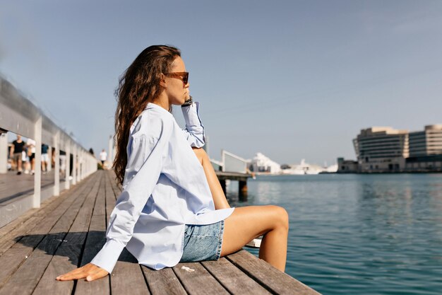 Profile portrait of stylish european girl in blue shirt and denim shorts is sitting on wooden pier and looking forward in sunlight on background of blue lake with yachts