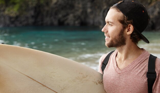 Profile portrait of surfer man in good mood, carrying his surfing board under his arm looking at sea and smiling, having thoughtful look