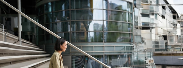 Foto gratuita il ritratto di profilo di una giovane donna asiatica con una studentessa portatile si siede sulle scale fuori dall'edificio e