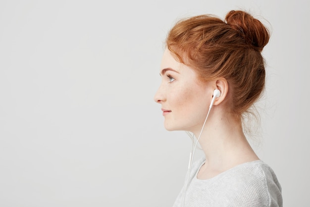 Profile portrait of young beautiful tender redhead girl with bun in headphones smiling .