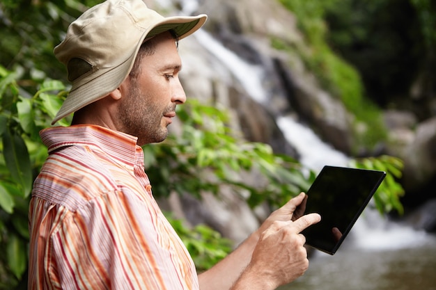 Profile of serious male scientist with stubble taking picture of nature on his black generic digital tablet while working on scientific research in jungle.