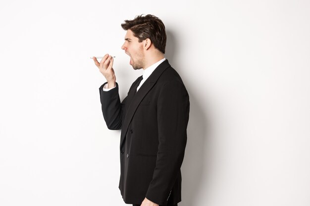 Снимок профиля разгневанного бизнесмена в черном костюме, кричащего по громкой связи и выглядящего безумным, записывающего голосовое сообщение, стоящего на белом фоне