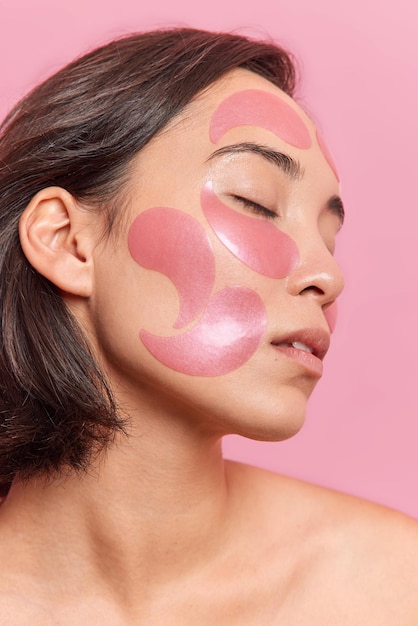 Profile shot of brunette asian woman keeps eyes closed applies hydrogel patches on face to refresh skin has healthy well cared skin dark short hair isolated over pink backgroud beauty concept