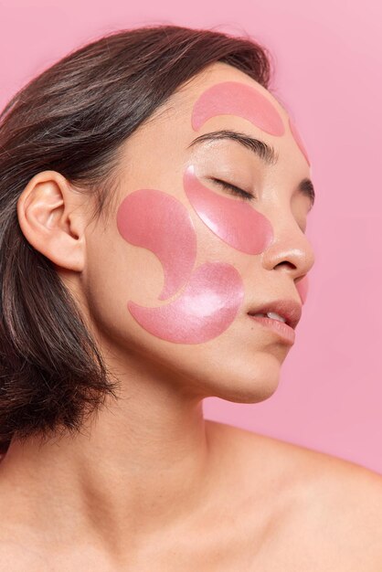 Profile shot of brunette Asian woman keeps eyes closed applies hydrogel patches on face to refresh skin has healthy well cared skin dark short hair isolated over pink backgroud Beauty concept