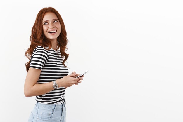 Снимок профиля беззаботная счастливая восторженная рыжеволосая женщина в полосатой футболке поворачивает налево и улыбается, радостно смеясь, держа смартфон, обмениваясь сообщениями, друзья подходят к стенду для вечеринок на белом фоне