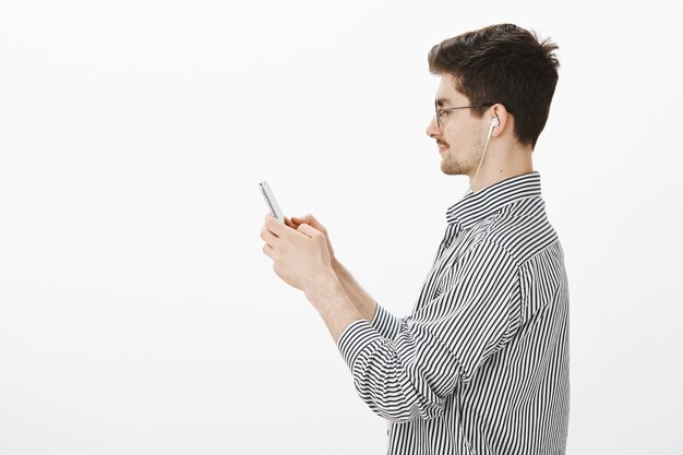 Profile shot of confident attractive bearded guy in trendy glasses and striped shirt, holding smartphone and looking at screen