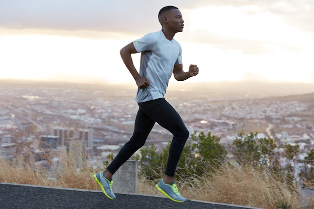 Free photo profile shot of energetic dark skinned man runs on highway against clear sky with morning light, wears casual comfortable t shirt, leggings and trainers, enjoys spare time at summer