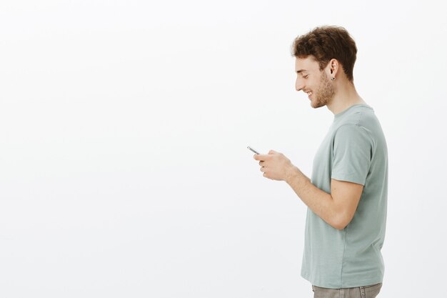 Profile shot of friendly handsome young male model in earrings, holding smartphone in hands, smiling broadly at screen, being satisfied with cute message from friend