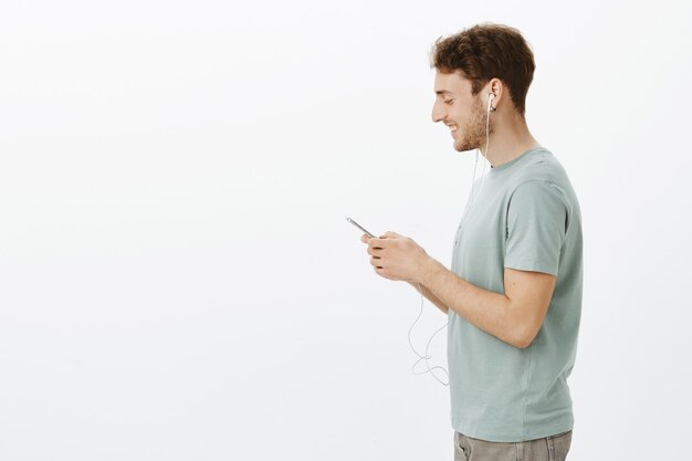 Profile shot of handsome joyful european male coworker with fair hair, holding smartphone and listening music in earphones