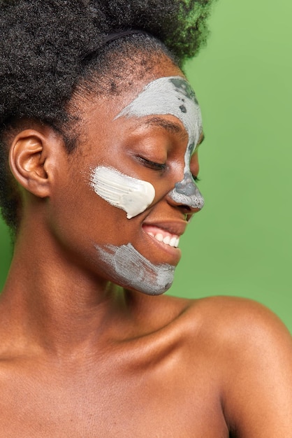 Profile shot of happy dark skinned woman applies clay facial mask for skin treatment keeps eyes closed stands shirtless has curly hair smiles gently isolated over green background Beauty procedures