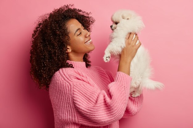 Profile shot of happy smiling woman raises miniature dog in both hands, looks with pleasure and smile, found stray pet, dressed in knitted jumper, poses over pink wall, expresses positive emotions