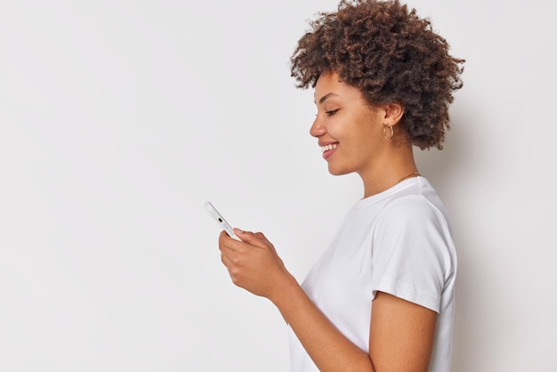 Profile shot of positive curly haired young woman uses mobile phone for communication messaging with important person dressed in casual t shirt isolated over white background blank copy space