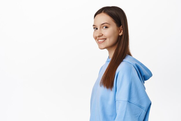 Снимок профиля молодой привлекательной женщины 20 лет, одетой в синюю повседневную толстовку с капюшоном, поворачивающейся лицом к камере и счастливой улыбающейся с белыми идеальными зубами, стоящей на белом фоне