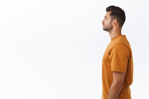 Profile studio shot attractive young modern hipster guy in brown stylish tshirt standing in line to buy coffee waiting someone as looking left casually no emotions standing white background