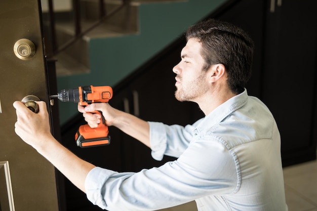 Profile view of an attractive handyman using a power drill to fix a door knob in a house