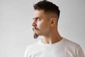 Free photo profile view of fierce displeased young caucasian male with goatee, mustache and stylish haircut poising isolated in white t-shirt looking away with angry offended expression, does not want to talk