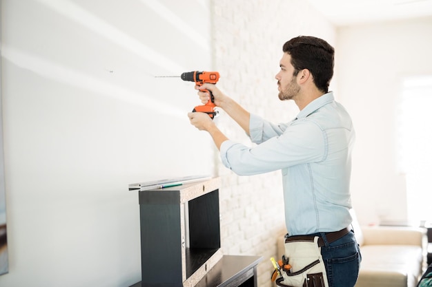 Profile view of a good looking young handyman making some drill holes in a wall and installing a shelf