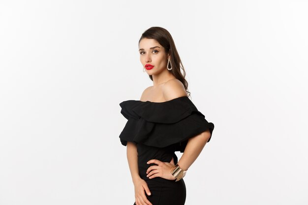 Profile view of sexy woman with red lips and makeup, wearing black dress, posing over white background.