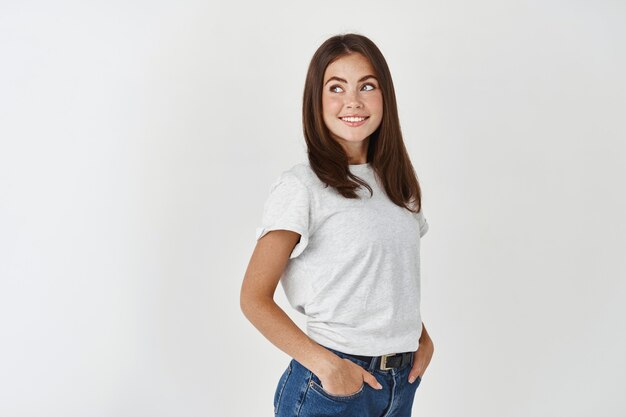 Profile of young ambitious woman looking left with interest, standing over white wall