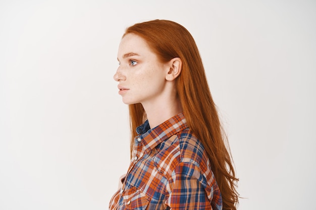 Profile of young redhead woman student with long natural ginger hair and pale skin, looking left, standing in casual shirt over white wall
