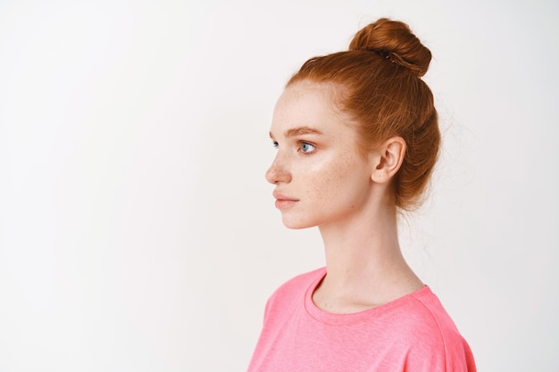 Profile of young woman with natural red hair combed in messy bun, looking left, standing over white wall. Facial treatment